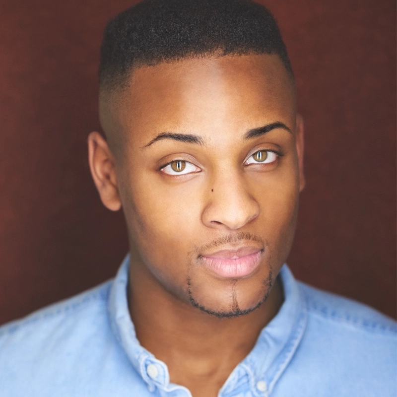Morgan Perry, a young man with light brown skin, hazel eyes, and a trimmed goatee, poses against a dark reddish-brown background. Wearing a light blue button-up shirt, he faces the camera with a neutral expression and closely-cropped hair.