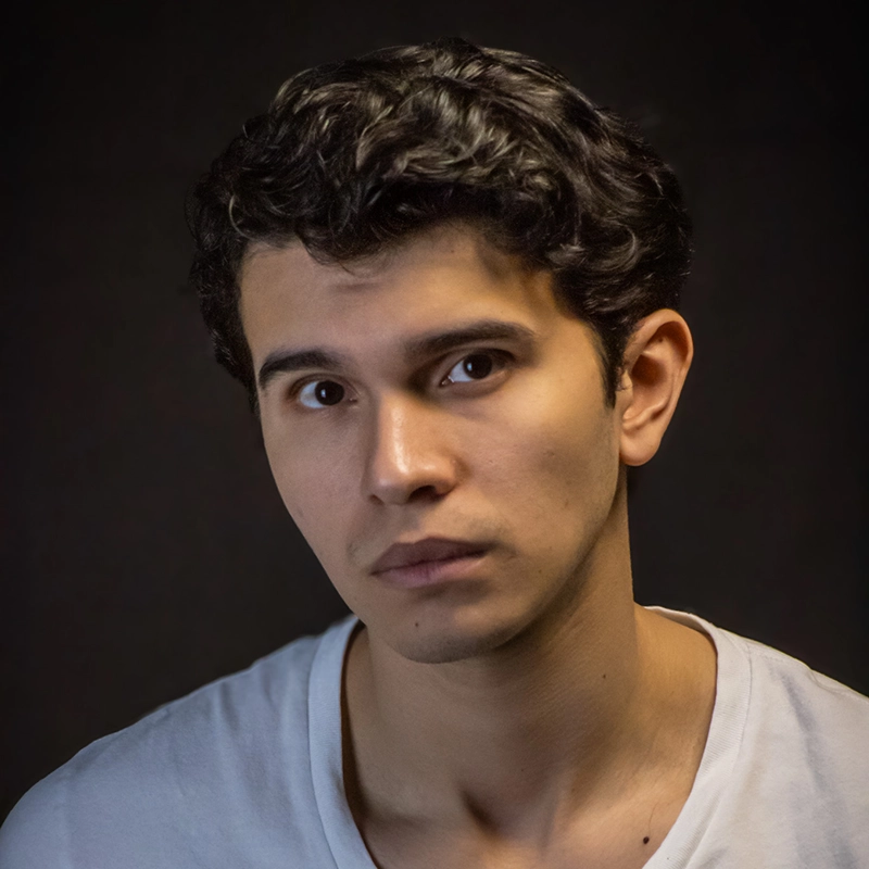 Mauricio Castillo, a young man with short, curly dark hair and light skin, looks directly at the camera with a neutral, slightly serious expression. He wears a light gray T-shirt against a dark background, the lighting highlighting his facial features.