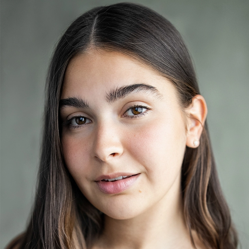 A young woman, Mayelah Barrera, with long, dark brown hair and clear skin looks slightly left of the camera, smiling softly. She has thick eyebrows, brown eyes, and wears small stud earrings. The background is neutral and softly blurred.
