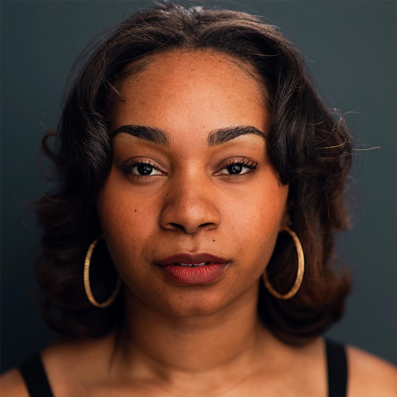 Morgan Perry, a young woman with medium brown skin and wavy, shoulder-length dark hair, looks directly at the camera. She wears gold hoop earrings and a black top, her calm, neutral expression set against a dark gray background.
