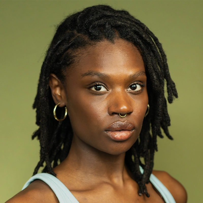 Morgan Perry, a woman with medium-dark skin and shoulder-length black locs, stares directly ahead. She wears gold hoop earrings, a gold septum ring, and a light blue top. The background is plain olive green; her expression is neutral and confident.