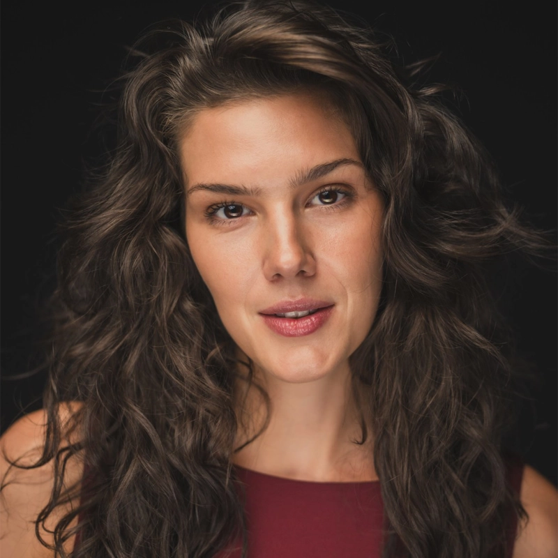 Christine Oczkus, with long, wavy brown hair and natural makeup, looks calmly at the camera. She wears a sleeveless dark red top, her features highlighted by soft, even lighting against a black background.