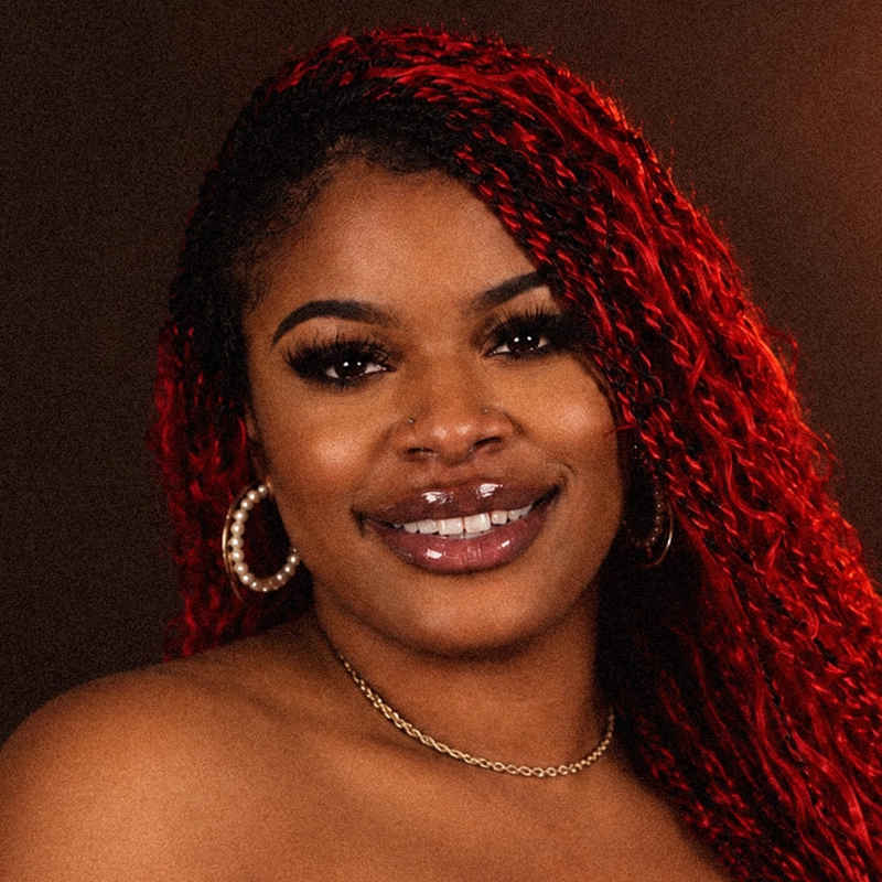 A woman with long, curly red hair, known as Shemaiah Red, smiles at the camera. She wears gold hoop earrings, a thin gold necklace, and glossy makeup. The dark background highlights her face, nose piercing, and visible eyelashes.