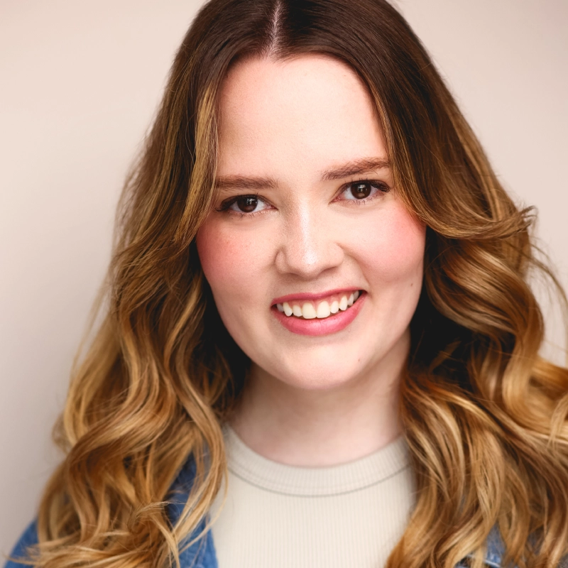 Katie Trumbull, a young woman with long, wavy blonde hair, smiles at the camera. She has fair skin, brown eyes, and wears a light top under a denim jacket against a soft beige background for a warm, inviting atmosphere.