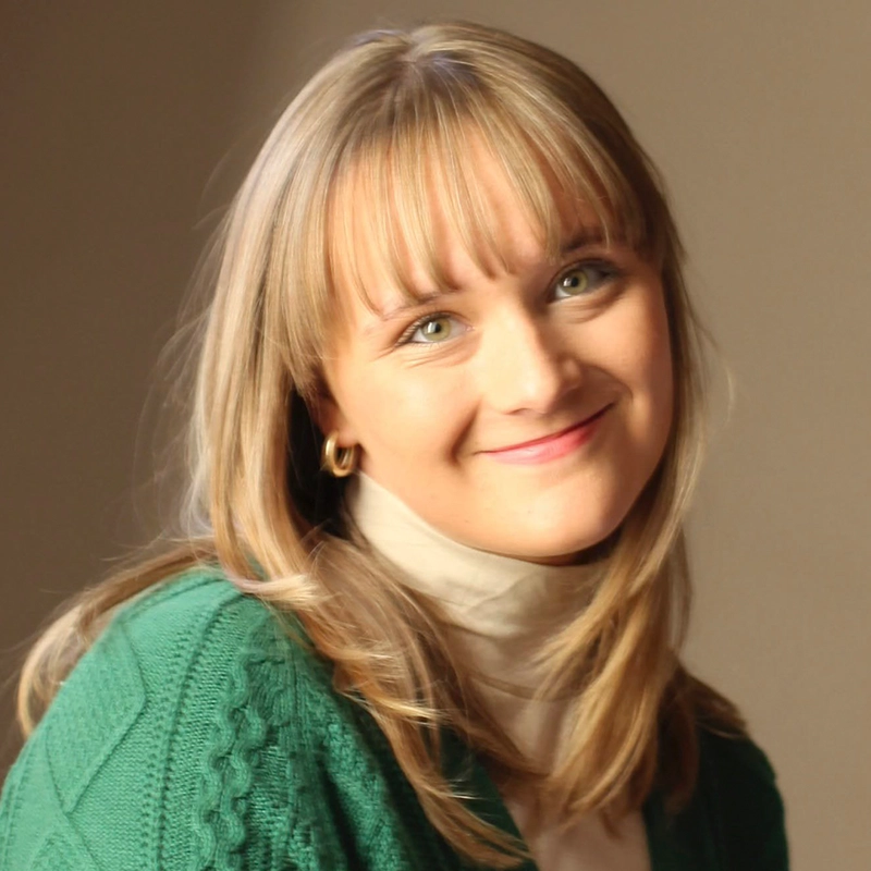 A young woman with blonde hair and bangs, wearing a green sweater over a light turtleneck, smiles gently at the camera against a plain beige background with soft lighting.
