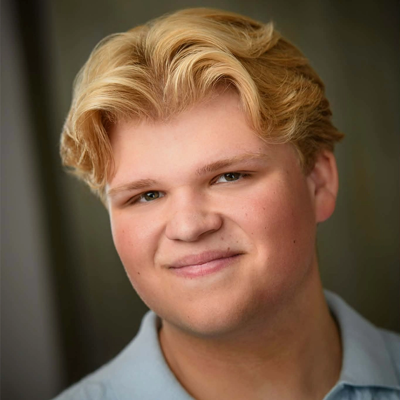 A young person with light skin and wavy blond hair smiles gently at the camera. Rockie Mosher wears a light blue collared shirt, posed against a softly blurred, neutral background. The lighting is even, highlighting their friendly expression.