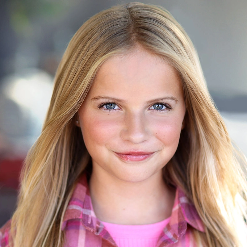A young girl with long blonde hair and blue eyes smiles gently at the camera. She is wearing a pink plaid shirt over a pink top. The softly blurred background brings focus to her face and expression, capturing Rockie Mosher’s youthful charm.