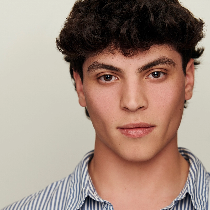 A young person with wavy dark hair and medium skin tone looks directly at the camera with a neutral expression. They wear a blue and white striped shirt against a plain, light-colored background.