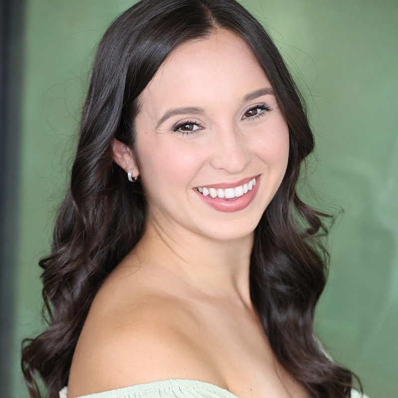 A young woman with long, wavy dark hair smiles brightly at the camera. She is wearing a light, off-the-shoulder top and small hoop earrings. The background is softly blurred in shades of green, creating a warm and welcoming atmosphere.