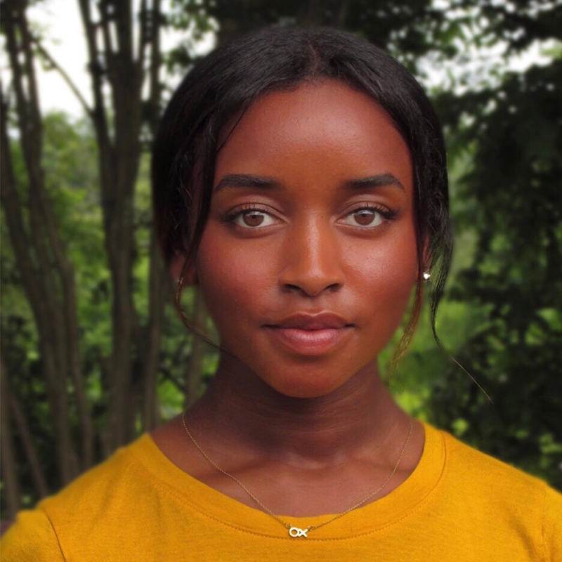A young woman with dark skin and straight dark hair, tied back with a few strands framing her face, stands outdoors. She wears a yellow top and a delicate necklace with an infinity symbol. The background shows green, leafy trees out of focus.