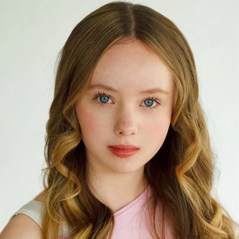 A young girl with long, wavy, light brown hair and blue eyes looks directly at the camera. She has fair skin, subtle makeup, and wears a pale pink top. The plain, light background keeps the focus on her face—evoking a Rockie Mosher portrait.