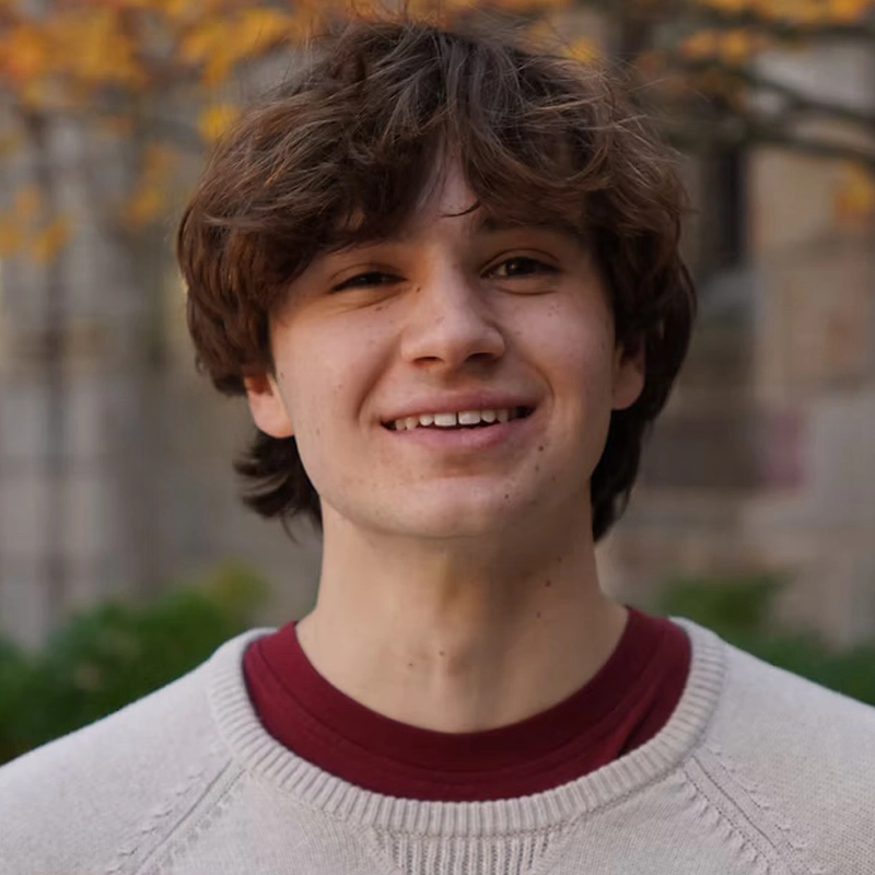 A young man with wavy brown hair smiles at the camera. He is wearing a cream-colored knit sweater over a maroon shirt. The background is outdoors with blurred autumn trees and building walls. The lighting is soft and natural.
