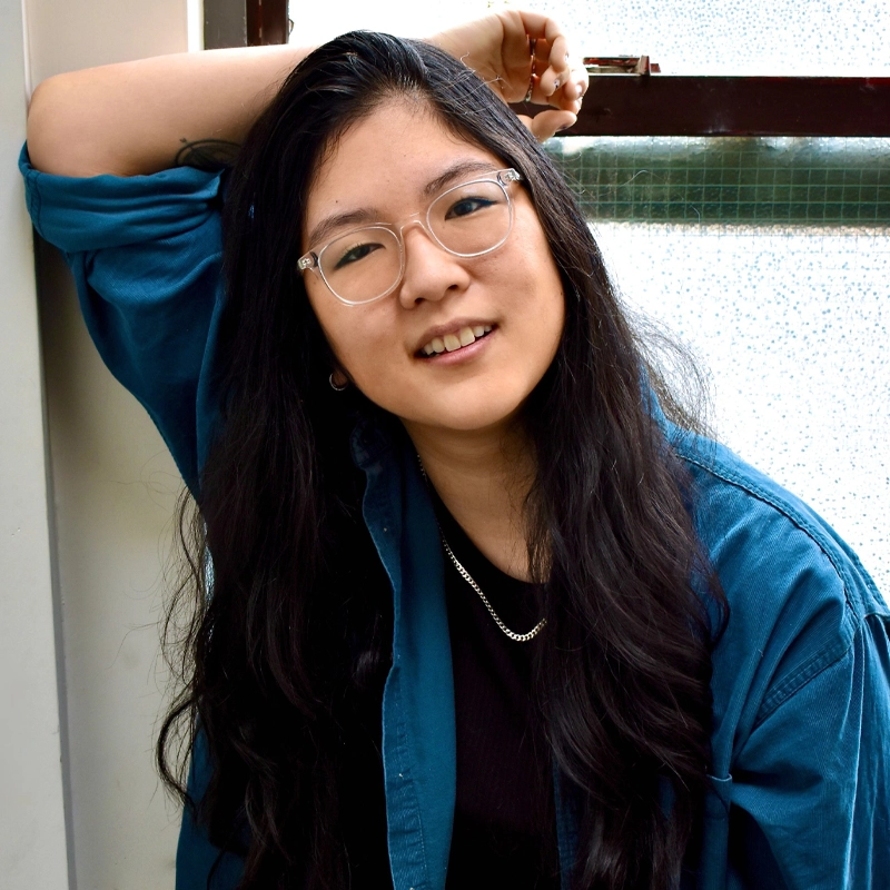 Mikayla Coxe, with long black hair, clear glasses, and a dark shirt under a blue button-up, leans against a window, smiling warmly. One arm is raised behind her head and a silver chain necklace shines softly in the gentle light.