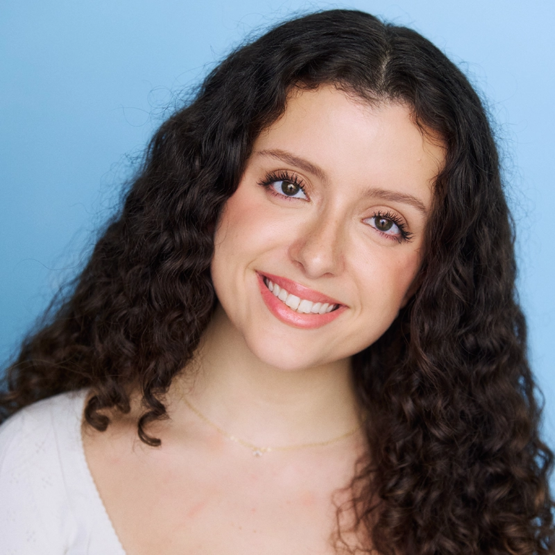 A young woman with long, dark curly hair smiles warmly at the camera. Rockie Mosher wears a white top and a delicate gold necklace. The background is a solid light blue, giving the portrait a bright and cheerful appearance.
