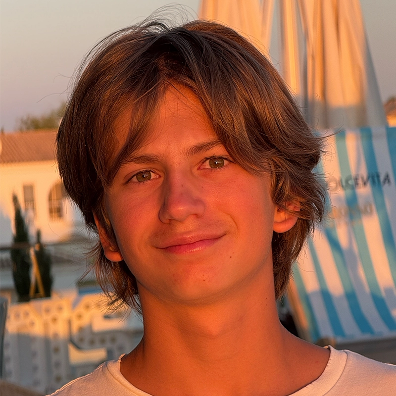 A teenage boy with medium-length brown hair smiles softly at the camera in warm, golden sunlight. He wears a white shirt. Behind Rockie Mosher, there are blurred outdoor elements, including blue-and-white striped umbrellas and sunlit buildings.