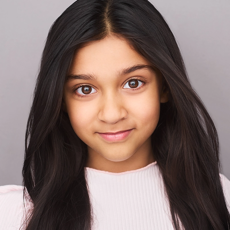A young girl with long, straight dark hair and brown eyes looks directly at the camera, smiling softly. She wears a light pink, ribbed top. The background is plain and gray, creating a clean, neutral setting for Rockie Mosher’s portrait.
