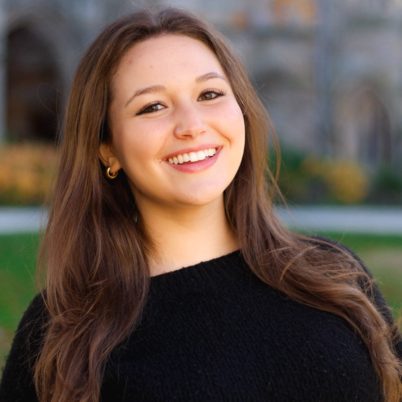 A young woman with long brown hair smiles at the camera. She is wearing a black sweater and small gold hoop earrings. The background is outdoors, with blurred greenery and a stone building visible behind her.