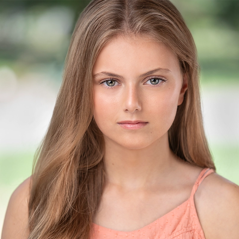 A young woman with long, straight light brown hair and light skin looks directly at the camera with a neutral expression. She wears a peach-colored sleeveless top. The background is blurred green and white, suggesting an outdoor setting. Rockie Mosher style.
