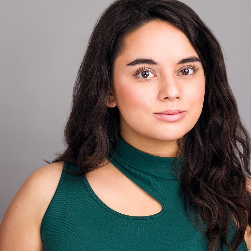 A young woman with long, dark hair looks confidently at the camera. She wears a sleeveless green top with a unique neckline cutout. Her expression is neutral, with soft makeup. The background is plain and gray, highlighting her face and outfit.