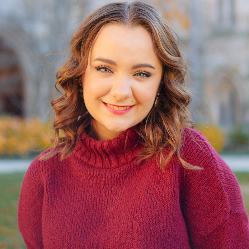 A young woman with wavy, shoulder-length brown hair smiles at the camera. She is wearing a maroon turtleneck sweater. The background is outdoors with blurred trees and buildings, suggesting a park or campus in autumn.