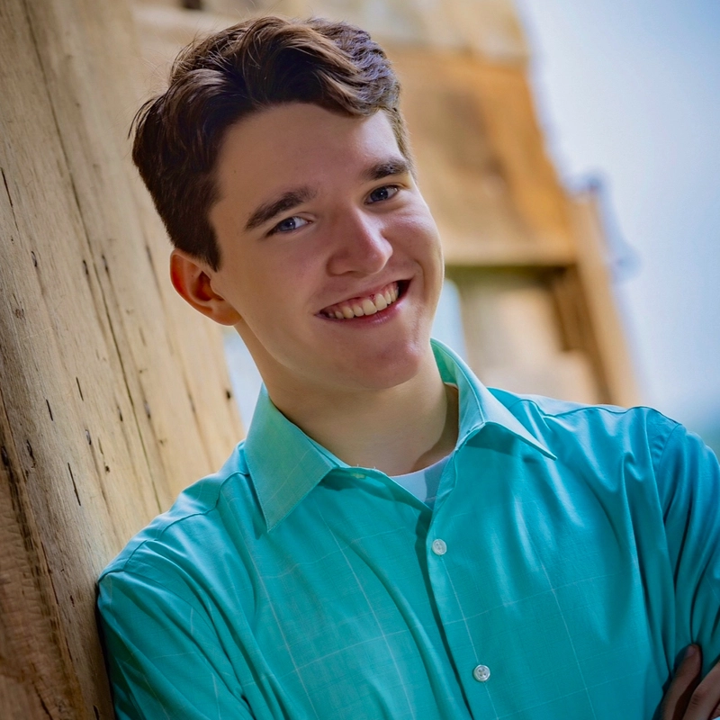 A young man with short brown hair smiles while leaning against a wooden wall. Dixon Miller wears a teal button-up shirt over a white undershirt. The background is blurred, suggesting an outdoor setting with natural light.