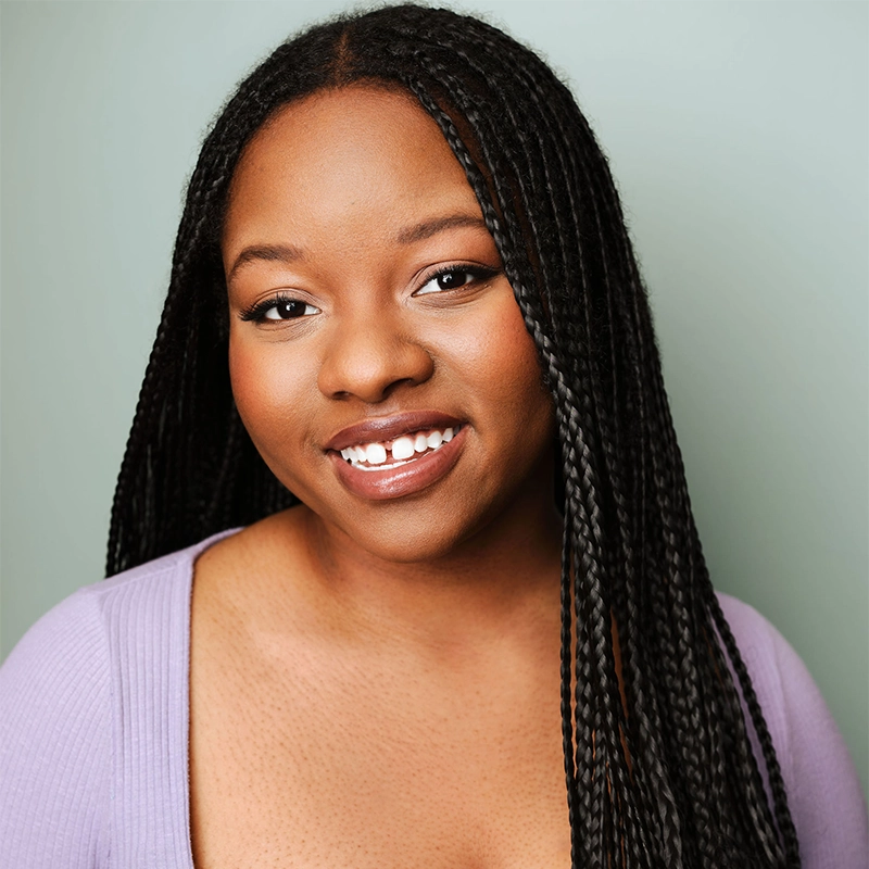 A woman with long, neatly styled braids smiles warmly. She wears a lavender top with a square neckline. Her skin is smooth, and her makeup is natural. The background is a soft, pale green, creating a calm and inviting atmosphere.