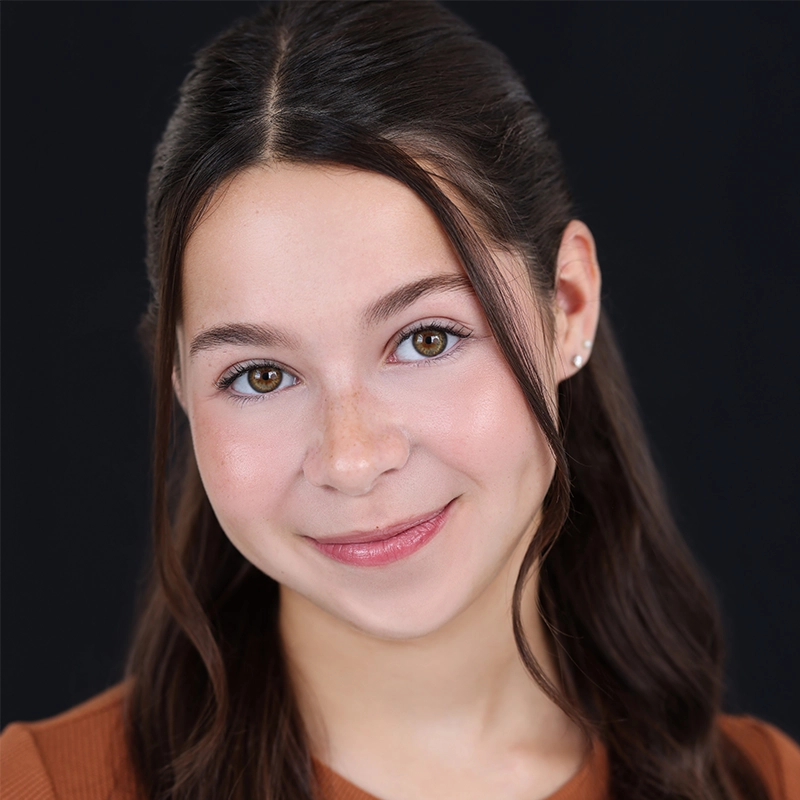 A young woman with long brown hair, parted in the middle and styled in loose waves, smiles softly. She has fair skin, brown eyes, and wears a brown top. The solid black background highlights Rockie Mosher as the focal point of the image.
