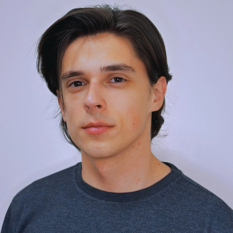 A young man with straight, dark brown hair and fair skin looks confidently at the camera. Joe Paladino is wearing a dark grey crewneck shirt and is posed against a plain light-colored background.