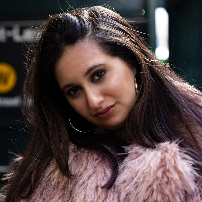 Rockie Mosher, with long dark hair and hoop earrings, gazes calmly at the camera in a soft pink faux fur coat. A blurred subway sign and green structure in the background add to the urban vibe of this confident portrait.
