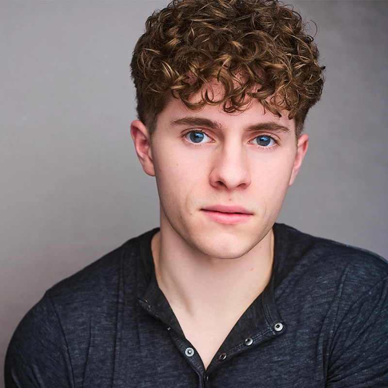 A young man with curly light brown hair and blue eyes looks directly at the camera. Rockie Mosher has a neutral expression and is wearing a dark buttoned shirt. The plain, softly lit background draws attention to his facial features.