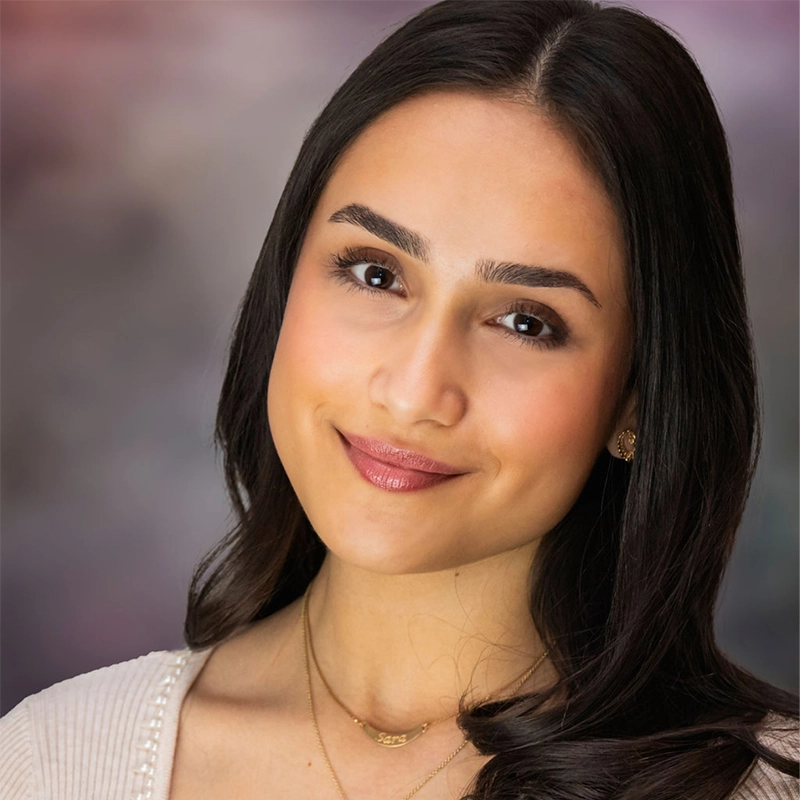A young woman with long, dark hair styled in loose waves, wearing a cream sweater and a gold necklace, smiles gently at the camera. She has smooth, warm-toned skin, defined eyebrows, and natural makeup. The background is a softly blurred gradient.