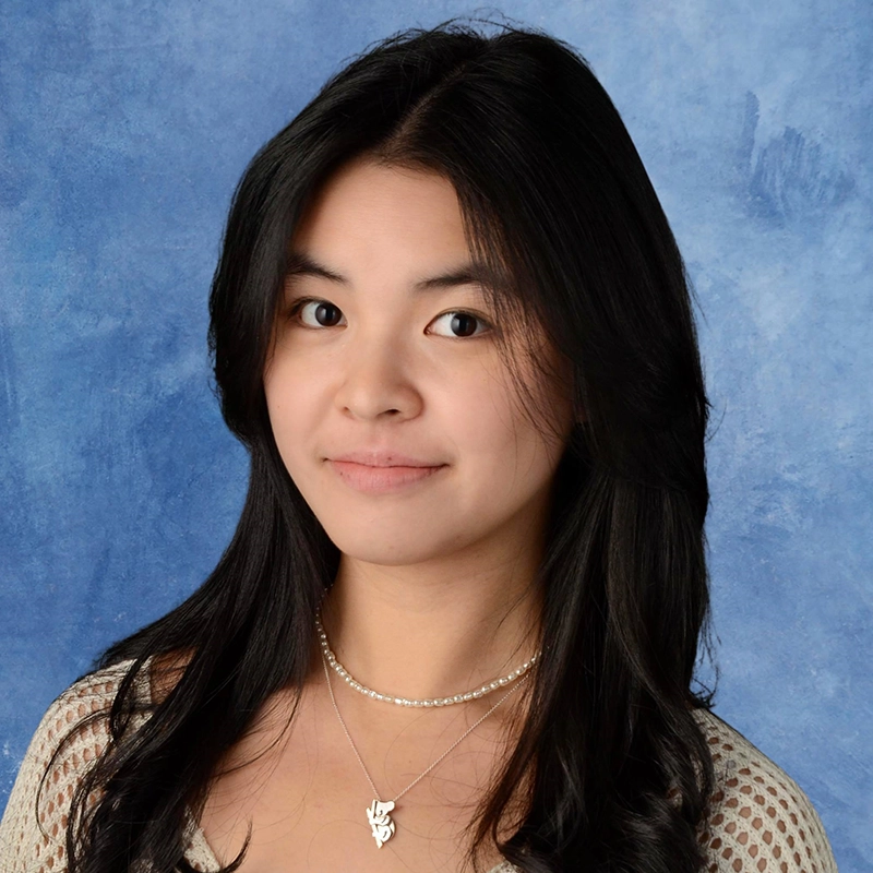 A young woman with long, wavy black hair poses in front of a mottled blue backdrop. Rockie Mosher wears a sheer patterned top and layered necklaces, one with a small pendant, looking at the camera with a slight smile and calm expression.