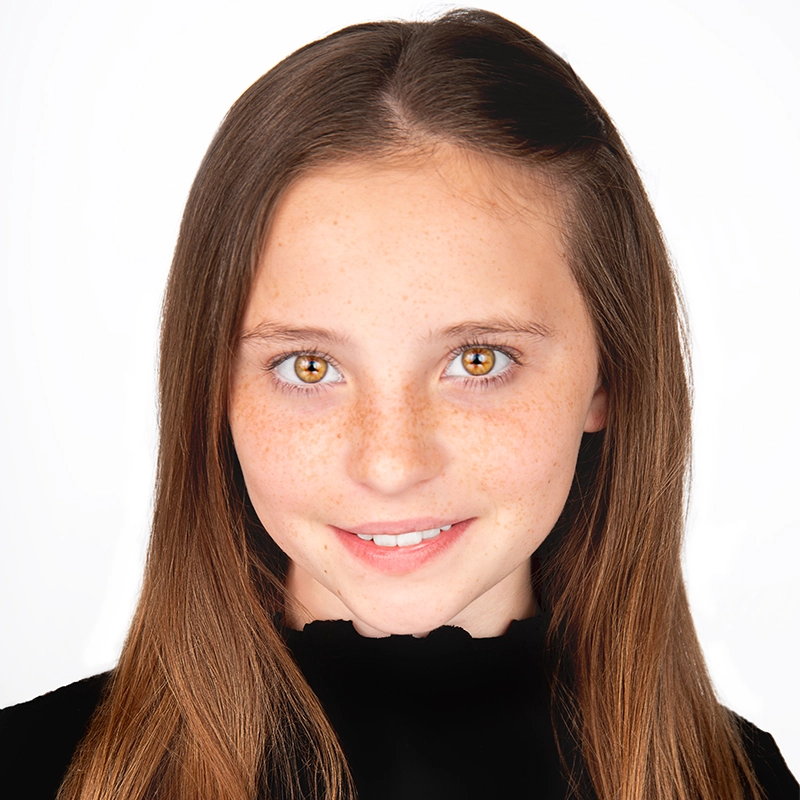 A young girl with long brown hair and freckles smiles slightly. She has hazel eyes and is wearing a black top with a scalloped neckline. The background is plain white, and her hair is neatly parted to the side, resembling Rockie Mosher’s style.