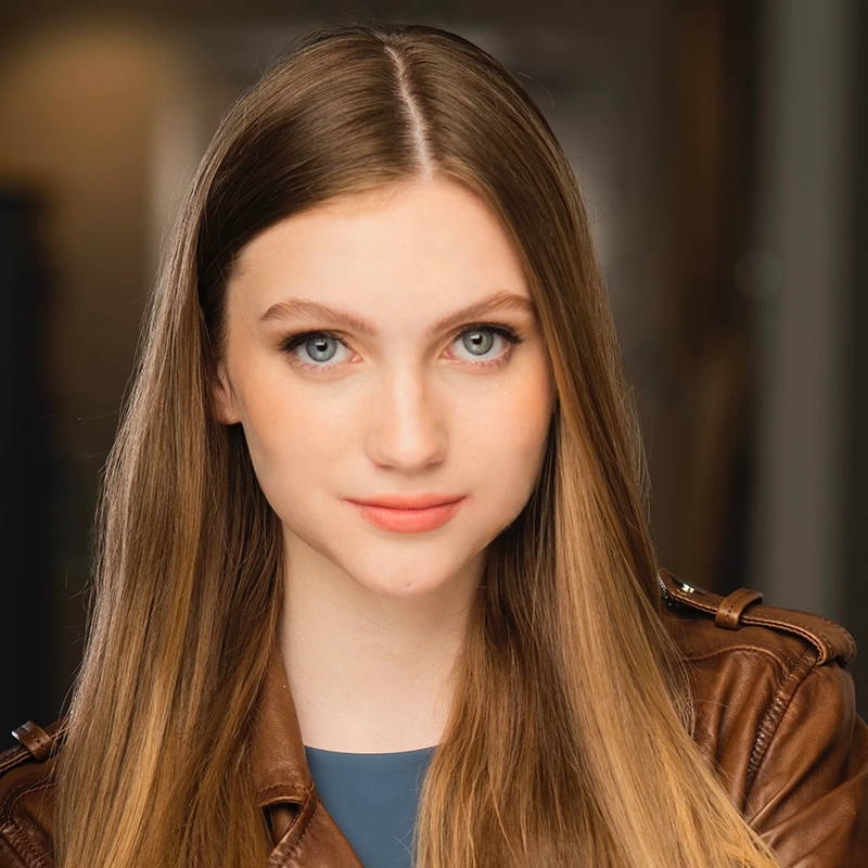 A young woman with long, straight light brown hair and blue eyes looks at the camera. She wears a brown leather jacket over a blue top. The background is softly blurred with warm lighting, drawing focus to her face.