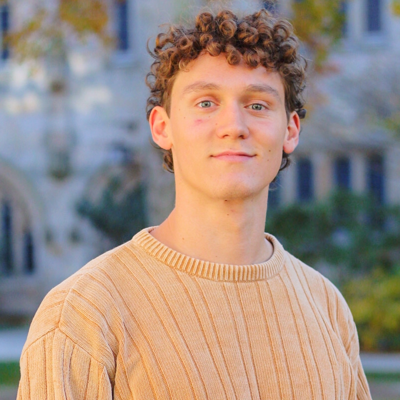A young person with short curly brown hair and blue eyes wears a beige ribbed sweater, standing outdoors with blurred trees and a stone building in the background. The lighting is bright and warm, suggesting a sunny autumn day—perhaps even August Taylor.