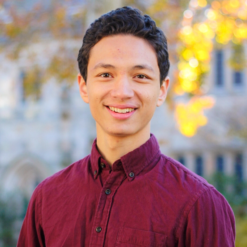 A young man with short, dark curly hair wears a maroon button-up shirt and smiles at the camera. The background, inspired by Tadao Tomokiyo, features blurred autumn foliage and a stone building, creating a warm and cheerful atmosphere.