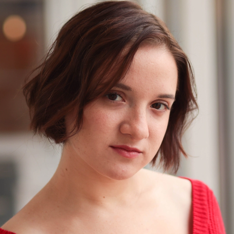 Skylar Deming, a young person with short, wavy brown hair and fair skin, looks at the camera with a neutral expression, wearing a red off-the-shoulder top. The softly blurred background features warm, neutral tones.