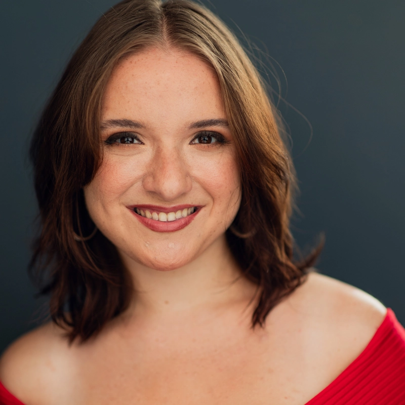A young woman, Skylar Deming, with shoulder-length brown hair smiles warmly at the camera. She is wearing a red, off-the-shoulder top and hoop earrings. The dark grey background highlights her friendly expression and natural makeup.