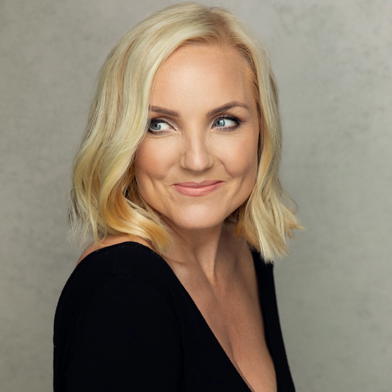 A woman with chin-length, wavy blonde hair and fair skin smiles softly while looking to her left. She wears a black V-neck top and is posed against a light gray, textured background. The lighting highlights her features and creates a gentle, professional look.