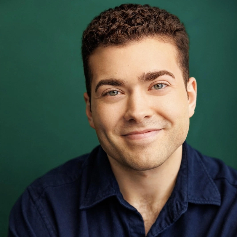 Alexander Fernandez, a young man with short, curly brown hair and light skin, smiles gently at the camera. Wearing a navy blue collared shirt, he sits in front of a solid green background with a friendly and approachable expression.