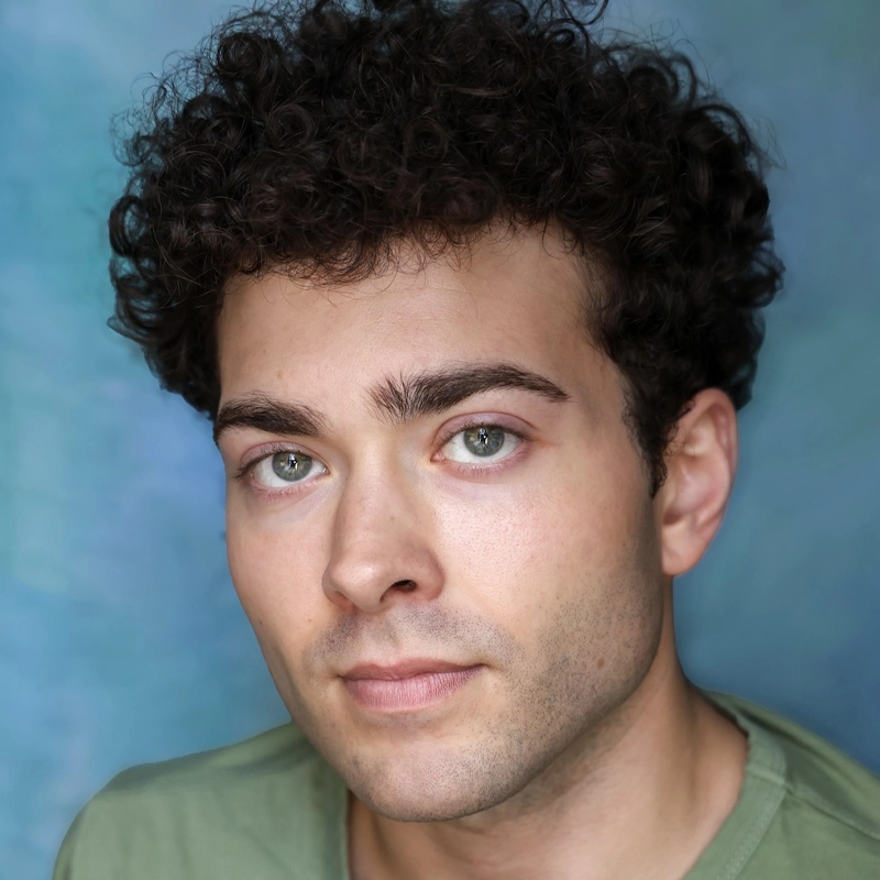 Young man, Alexander Fernandez, with short, dark curly hair and light eyes looks directly at the camera with a neutral expression. He has fair skin and is wearing a green shirt. The background is a mottled blue and green. The lighting is soft and even.