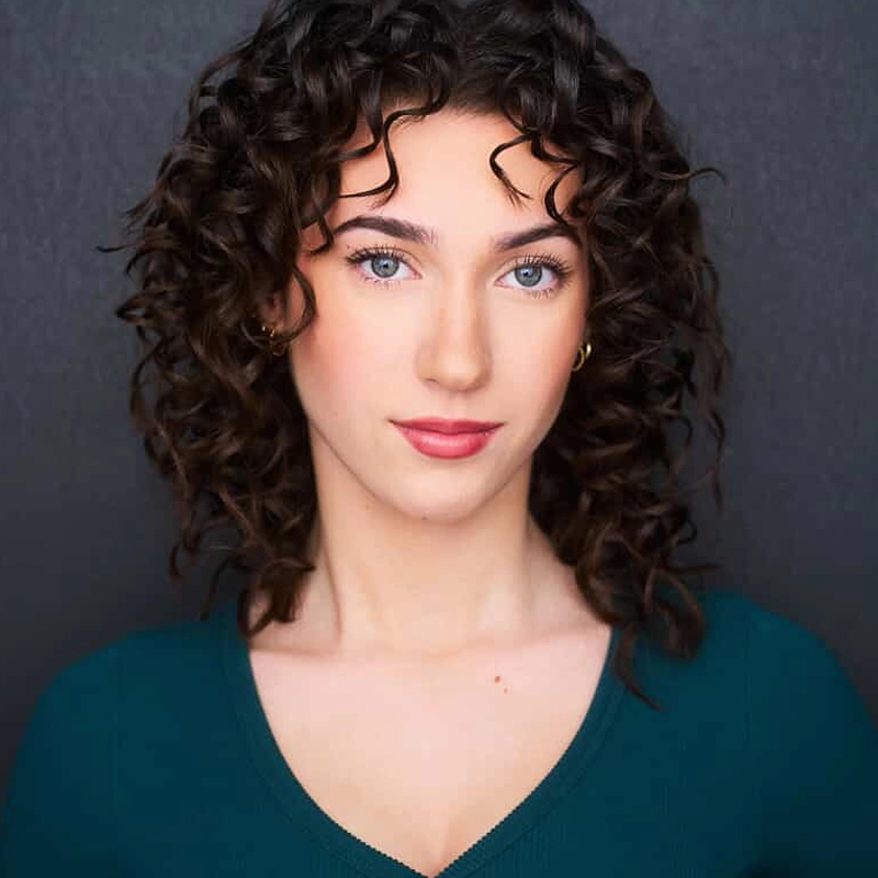 Olivia Flaherty, with fair skin, curly dark brown hair, and blue eyes, looks directly at the camera. She wears a teal V-neck top and small hoop earrings, her expression neutral against a dark, plain background.