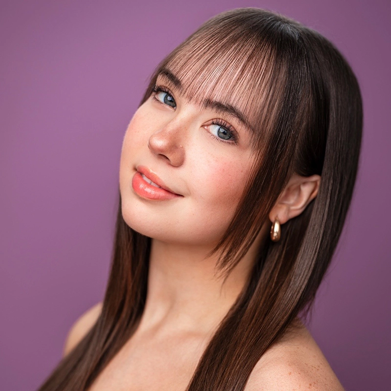 A young woman, Lauren Hartley, with straight brown hair and wispy bangs smiles softly. She has blue eyes, peach lipstick, and gold hoop earrings. Her complexion is fair with subtle freckles. The background is a smooth, solid purple.