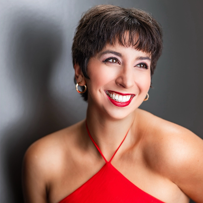 A woman with short dark hair smiles brightly, showing teeth. She wears a sleeveless red halter dress and gold hoop earrings. Her bold makeup features red lipstick. The portrait, photographed by Julian Lokash, has a smooth gray background with a soft shadow.