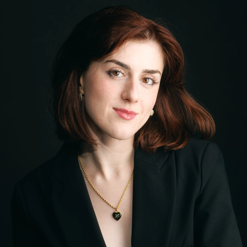 A woman with shoulder-length auburn hair, identified as Whitley Armstrong, is wearing a black blazer. She has on a gold heart-shaped necklace and small hoop earrings. The dark background contrasts with her fair complexion. Her expression is calm as she looks directly at the camera.