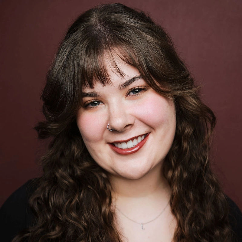 A young woman with long curly brown hair and bangs smiles at the camera. She has fair skin, a nose ring, and wears natural makeup with peachy blush and red lipstick. Lauren Hartley is dressed in a black top and a delicate necklace. The background is solid maroon.