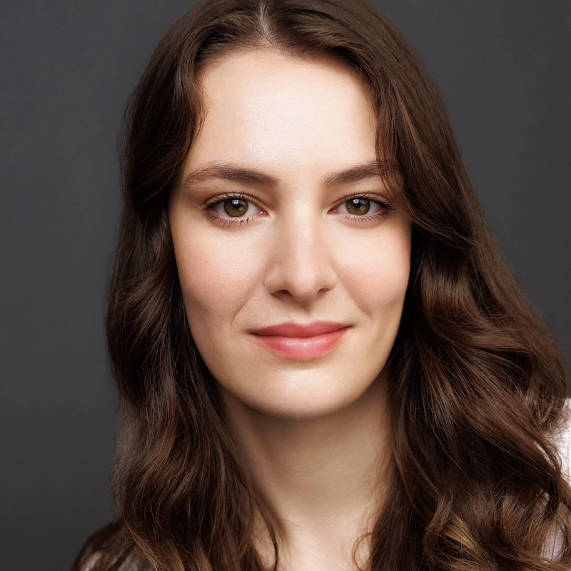 A young woman with long, wavy brown hair and fair skin smiles softly. Reminiscent of Lizzie J. Maguire, she has brown eyes, natural makeup, and wears a white top. The background is plain dark gray, giving the portrait a professional, studio-lit appearance.