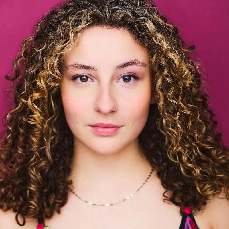 Sophia Anna O’Brien, a young woman with fair skin and brown eyes, faces the camera against a magenta background. She has shoulder-length, curly brown hair with blonde highlights and wears a gold necklace and a floral-patterned top.