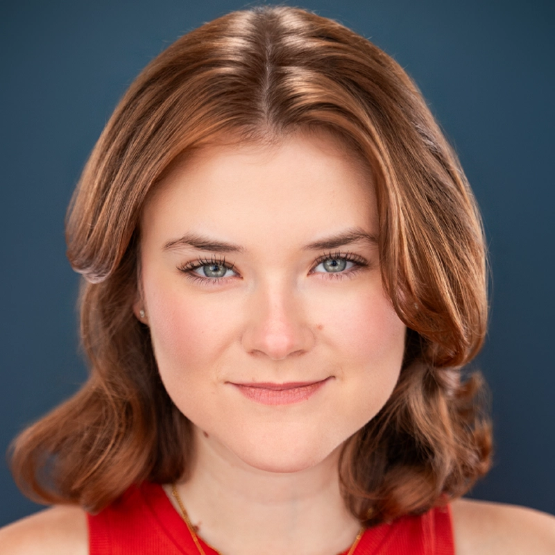 A woman with fair skin and shoulder-length light brown hair styled in soft waves, wearing a sleeveless red top. She has blue eyes, a slight smile, and is facing forward against a solid dark blue background.