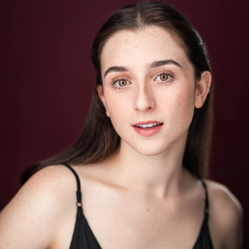 A young woman with fair skin, brown eyes, and long dark hair looks into the camera. She wears a black spaghetti strap top and has a gentle, natural smile. The background is a solid dark red. The lighting is soft, highlighting her clear complexion, giving Gemma Strauss an effortlessly elegant presence.